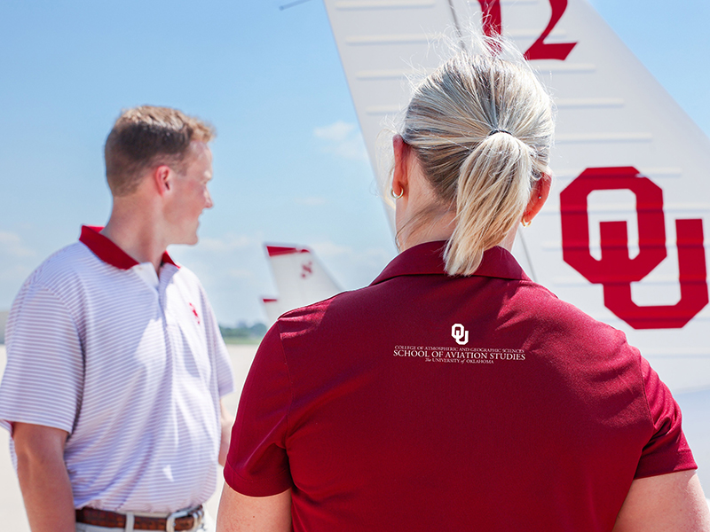 Two people talking with the tail of an OU airplane in the background.