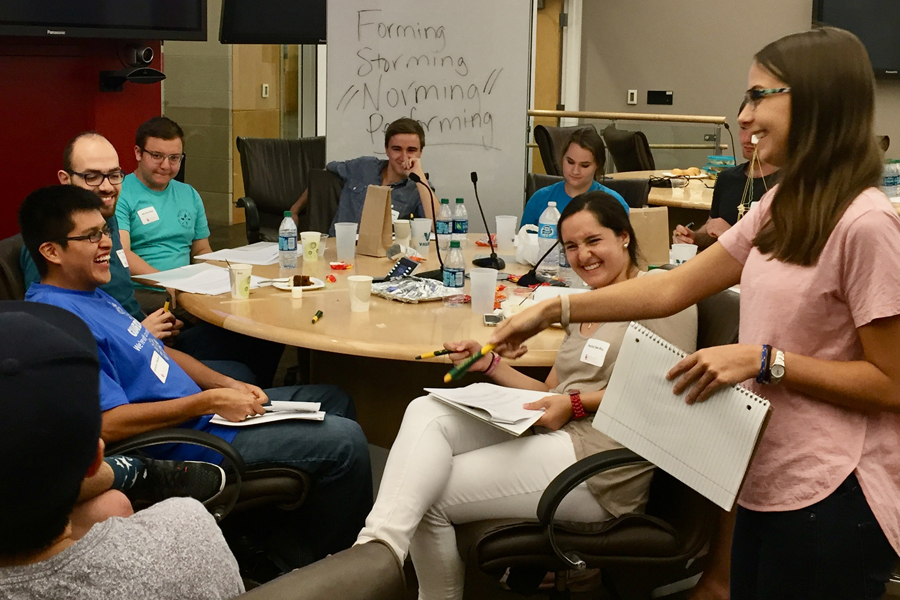 Six students sitting around a round table with one student standing and smiling.