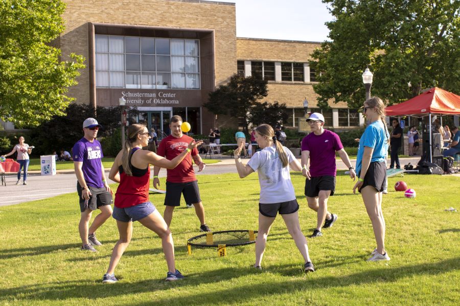 Group of students outside hitting a small yellow ball between each other. There is a building with the words Schusterman Center University of Oklahoma in the background.