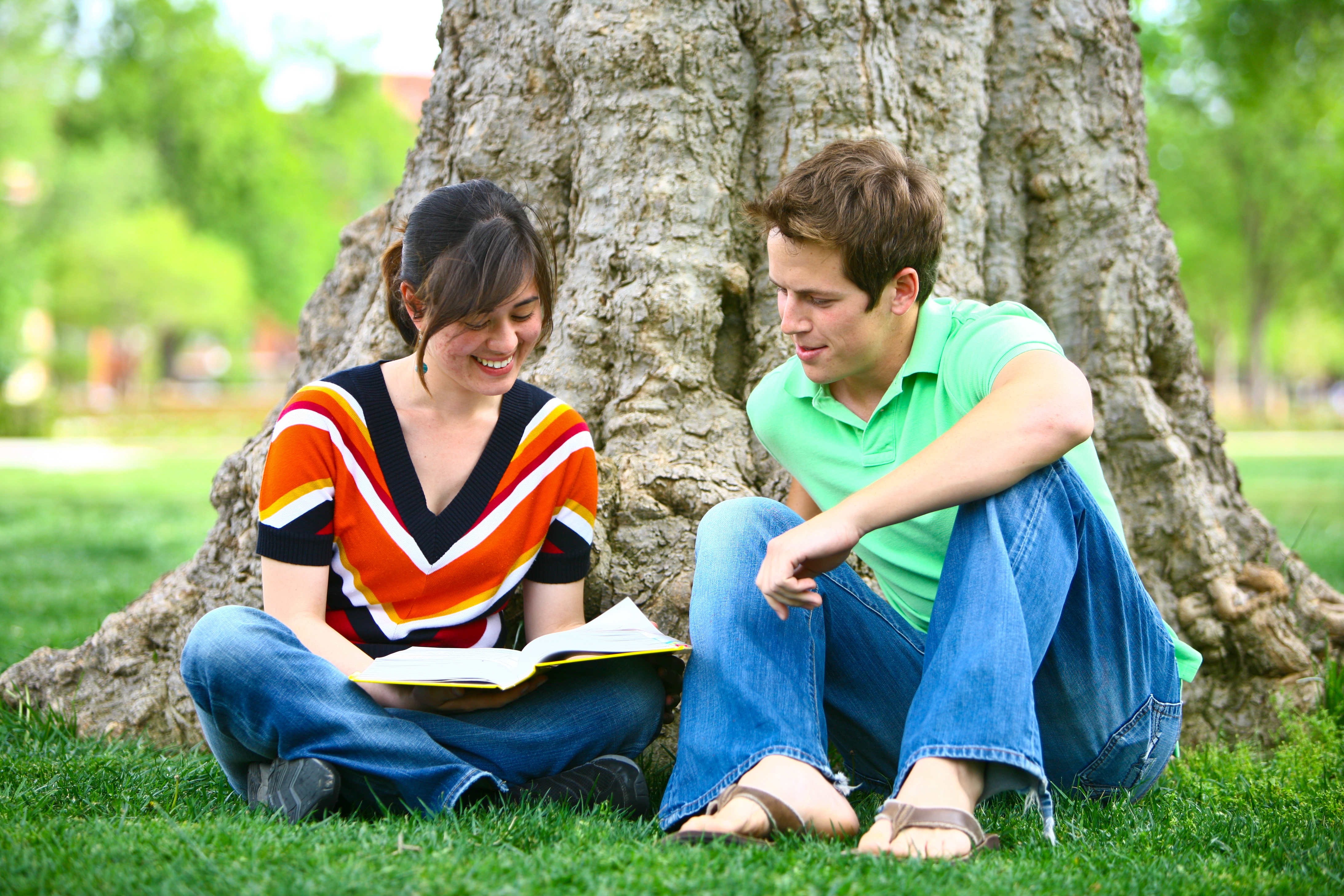 A classroom with students seated at small individual desks, each with either papers or phones on their desks. In the first row, three students are smiling and talking to each other. The student in the middle is writing something on a piece of paper