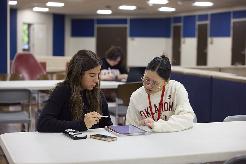 A classroom with students seated at small individual desks, each with either papers or phones on their desks. In the first row, three students are smiling and talking to each other. The student in the middle is writing something on a piece of paper