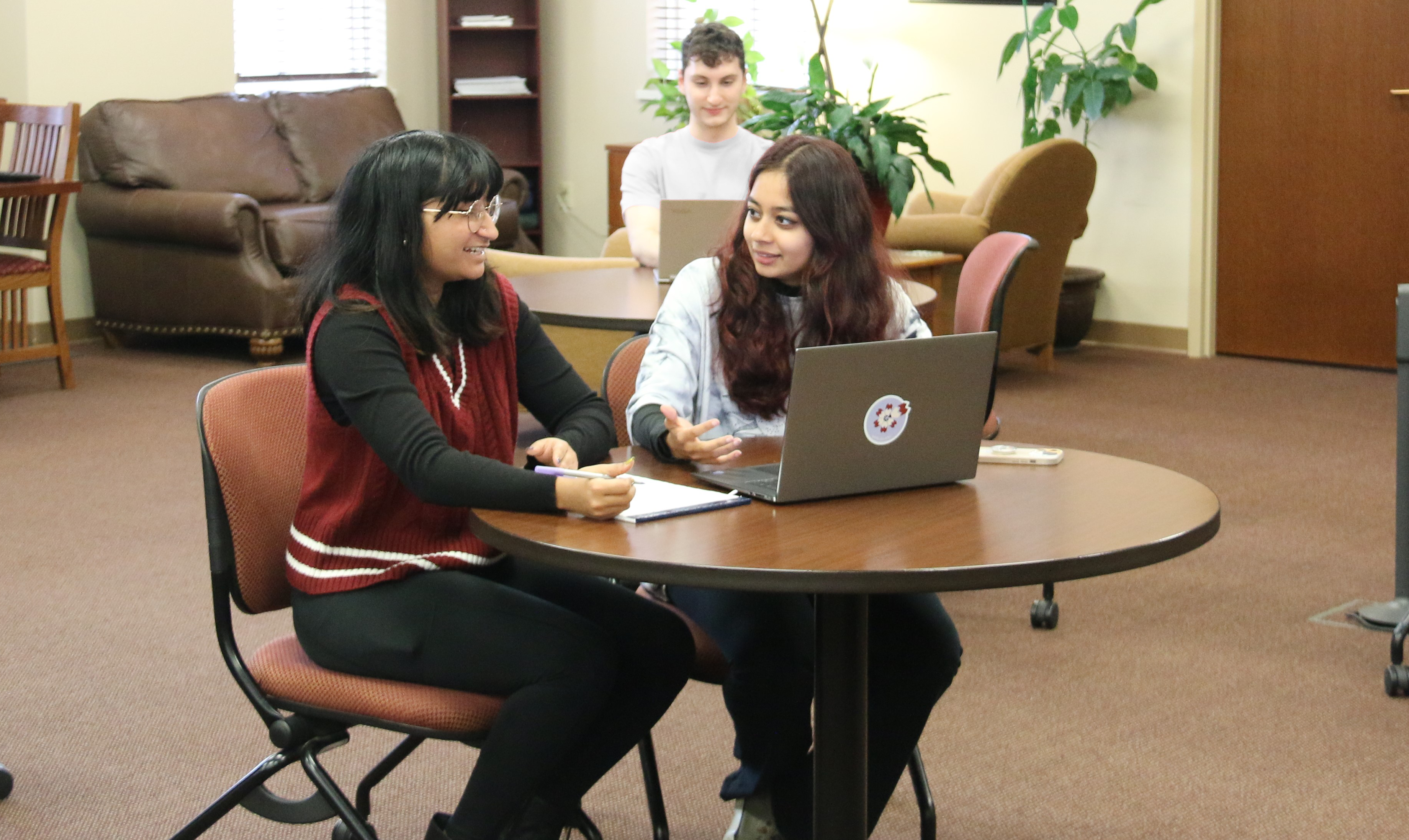 Two student sitting at a table, writing notes and reading a book.