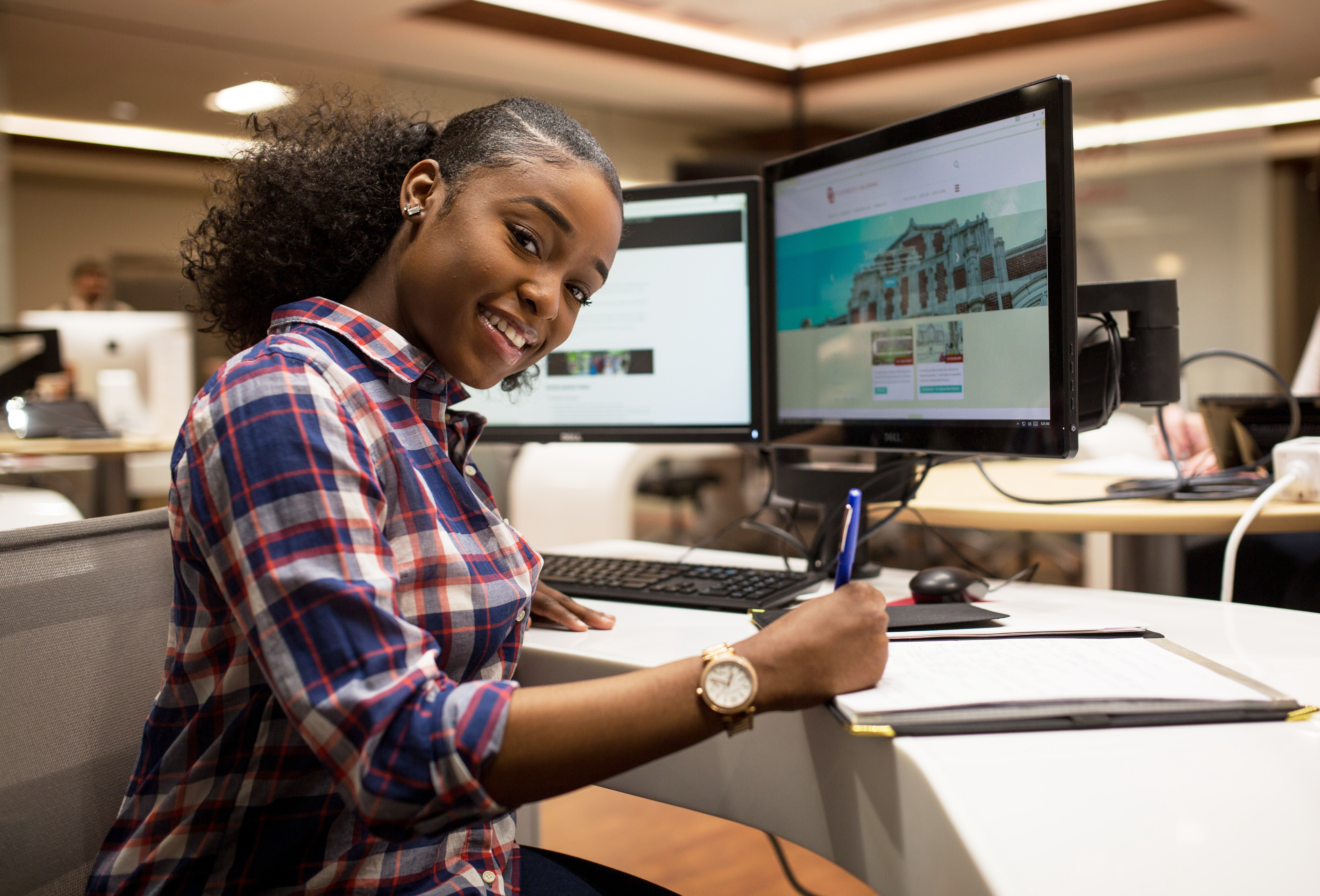 A woman sitting at a desktop computer with a pen in hand.