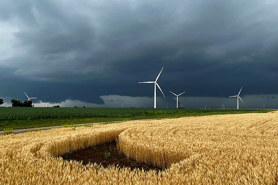 A broadcast meteorologist delivers a forecast in front of a green screen and surrounded by people at laptops.