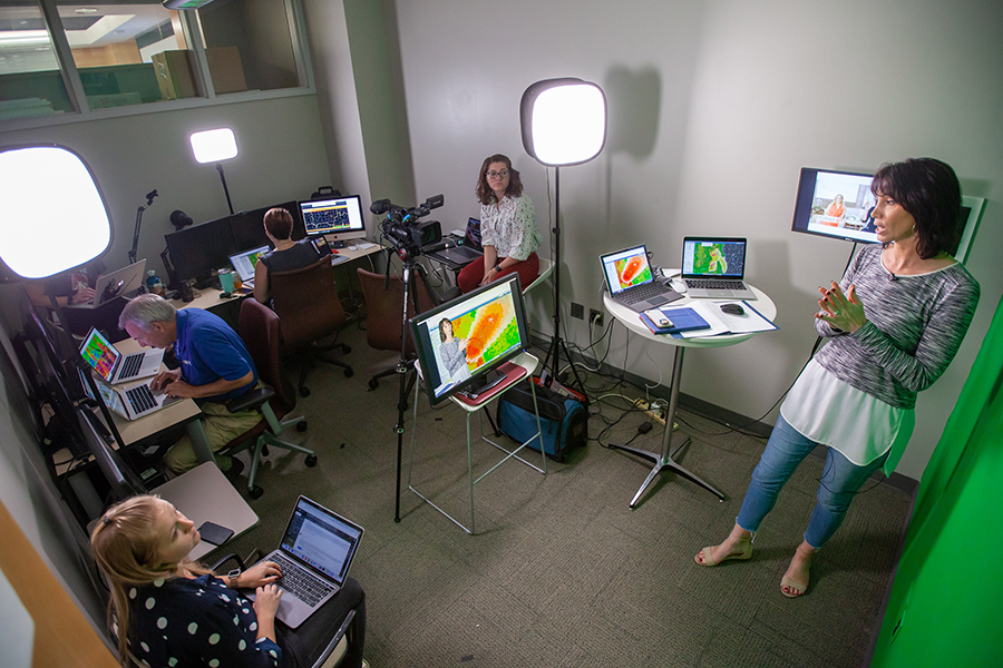 A broadcast meteorologist delivers a forecast in front of a green screen and surrounded by people at laptops.