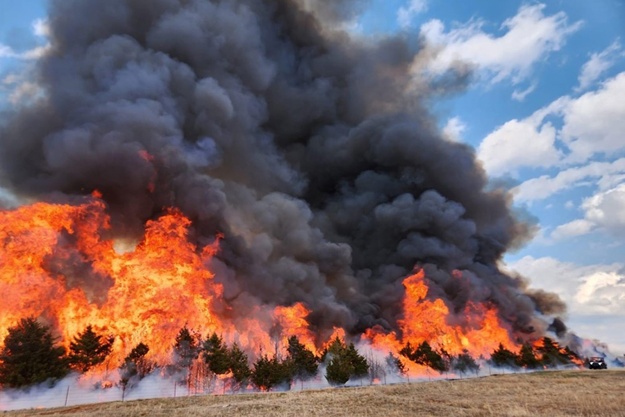 A fire rages across a landscape, with billowing gray plumes of smoke ascending into the sky.