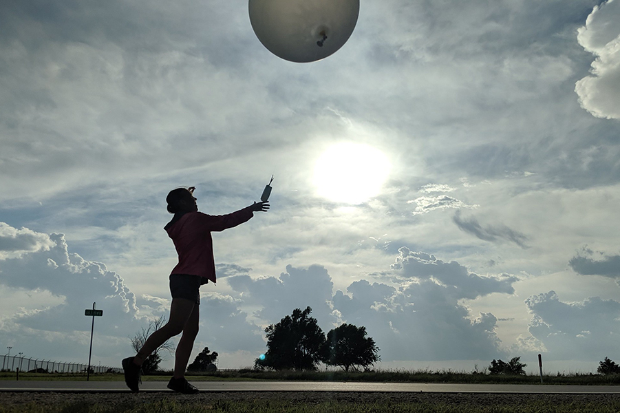 A silhouette of a person releases a radiosonde and weather balloon into a mostly cloudy sky.