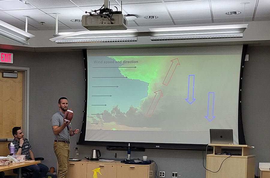 A scientist holds a football against a projected presentation to describe wind sheer.