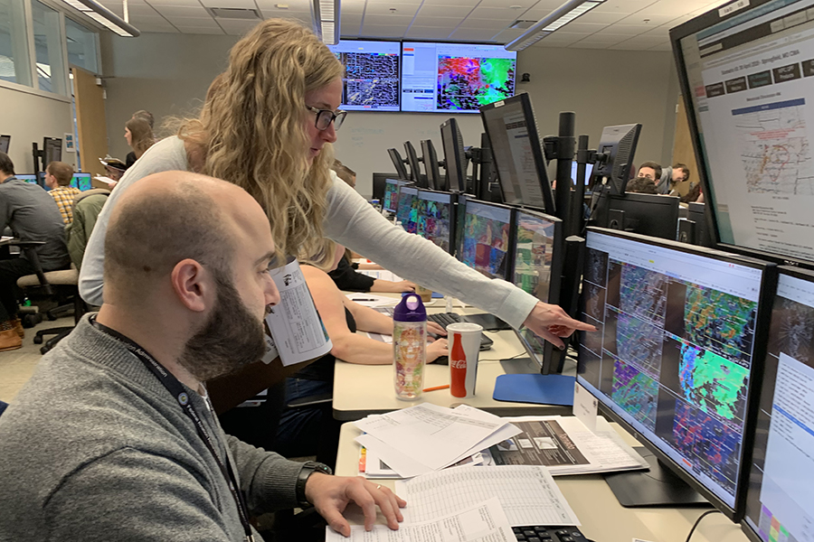An instructor points to a monitor displaying radar images while a forecaster looks on
