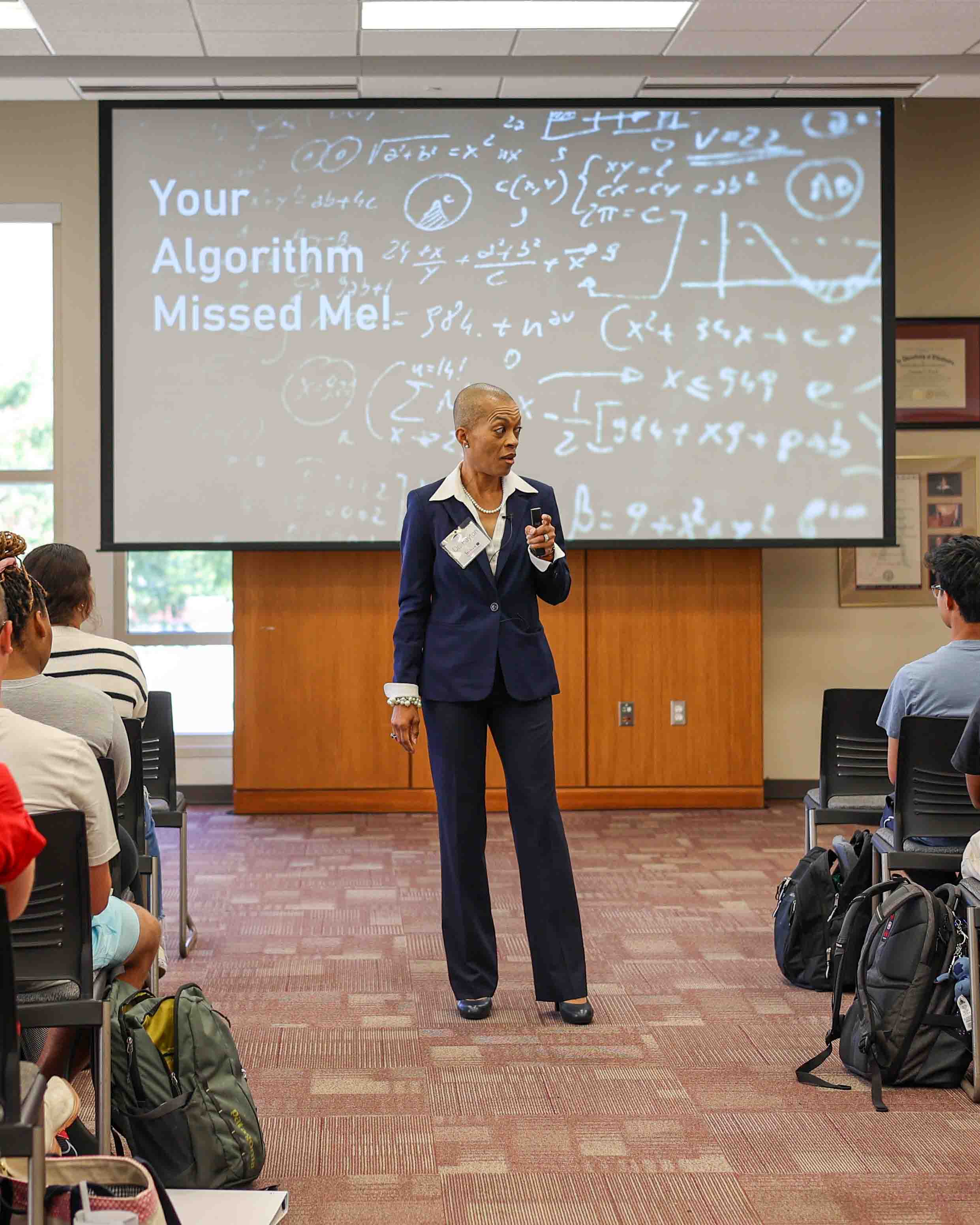 A woman speaks in front of a screen with the words "Your Algorithm Missed Me!"