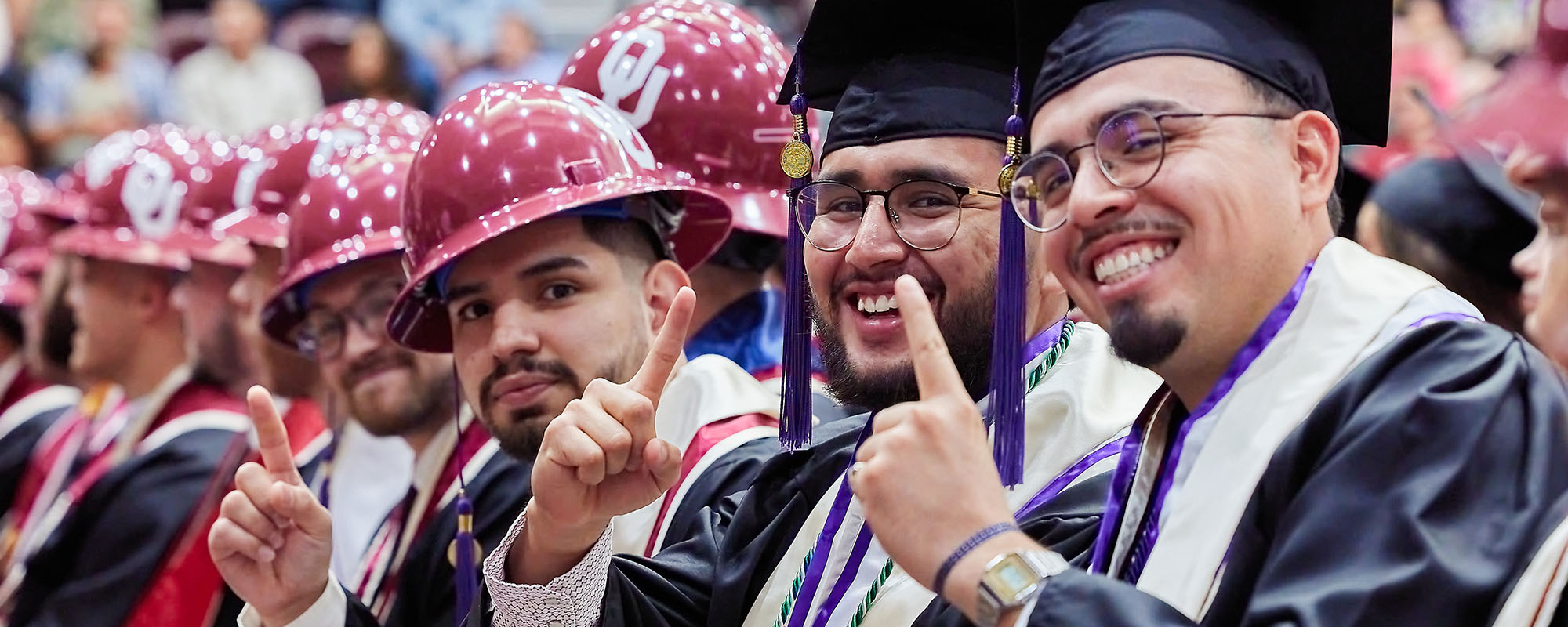 Students wearing hard hats and graduation regalia, holding up their pointer fingers.