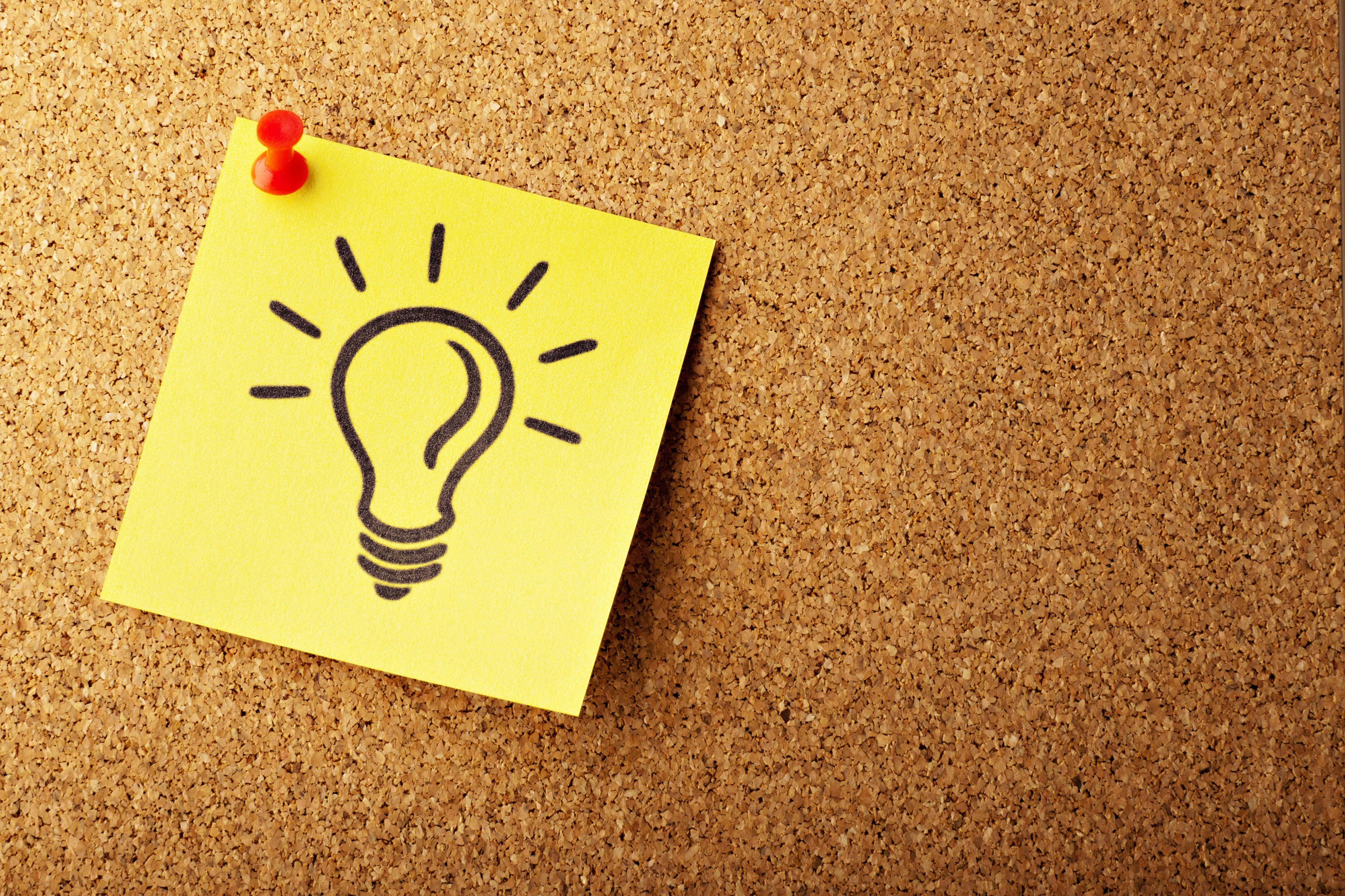 A yellow sticky note pinned to a cork bulletin board with a red pushpin, featuring a hand-drawn black light bulb icon 