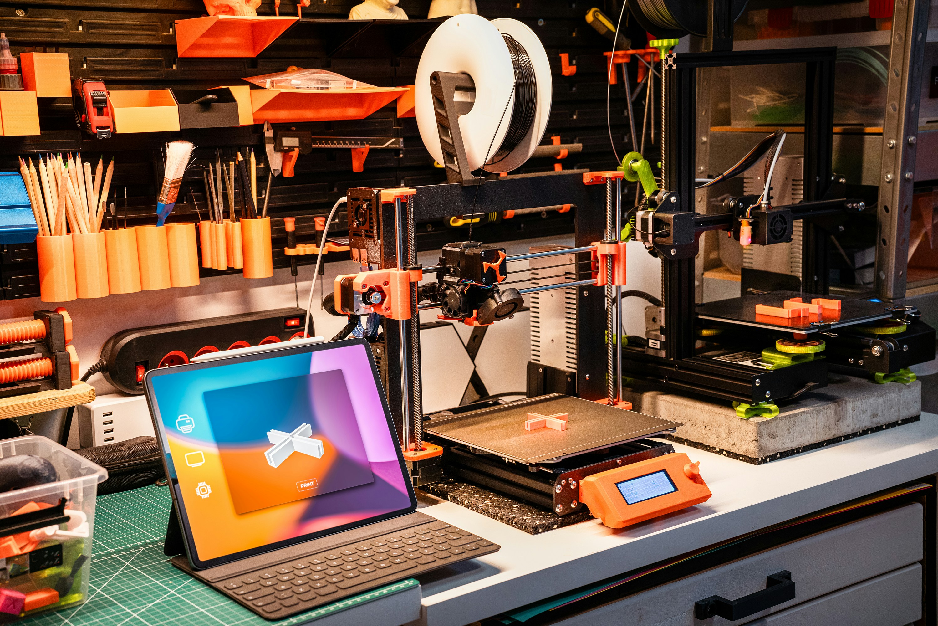 3D printing workshop with a tablet displaying a software, and orange-accented wall-mounted tool storage holding spools, pencils, and verious supplies