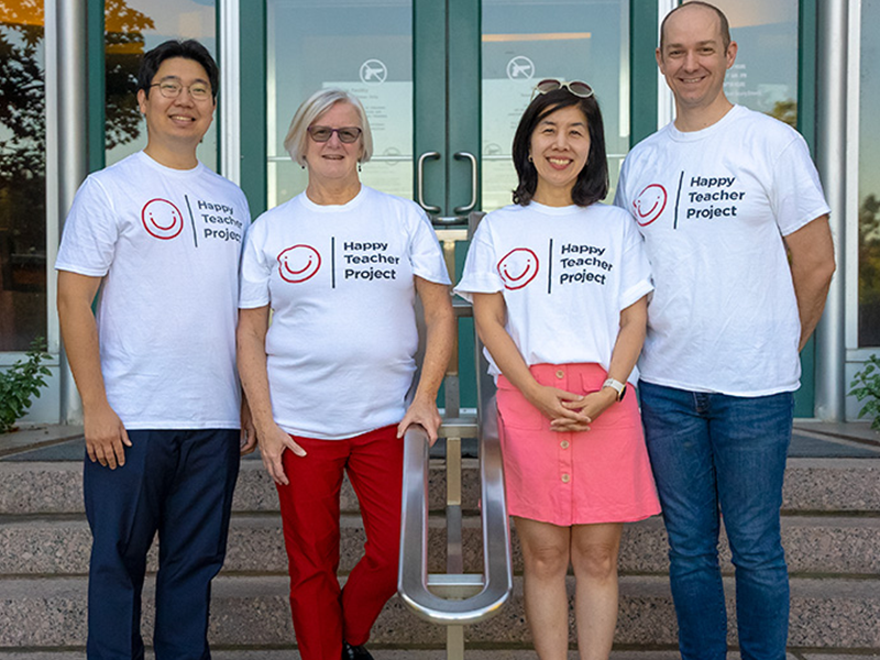 four people smiling on a staircase. All four are wearing the same white shirt that says "Happy Teacher Project"