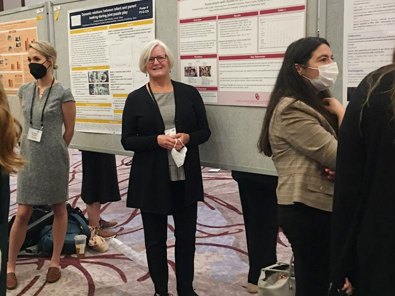 A woman in a black pantsuit with short gray hair standing in front of a bulletin board with a research poster taped to it.
