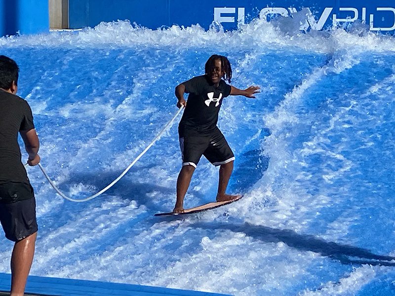 Student wearing black shorts and shirt standing on a surf board holding a rope.