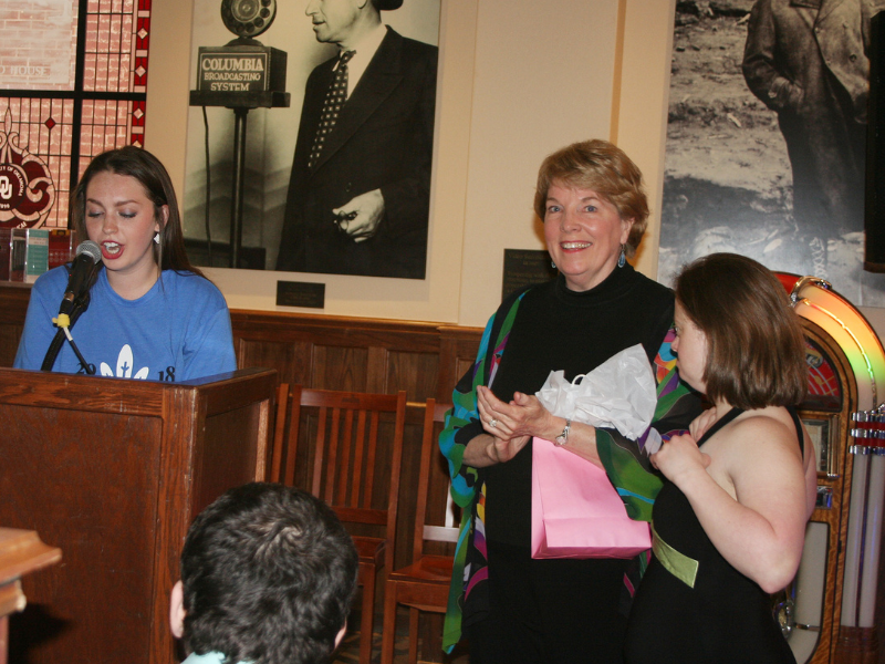 Woman talking at podium standing next to a woman in a black dress with a third woman standing next to her