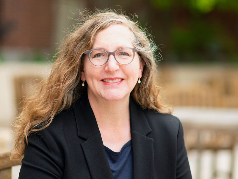 Woman with long hair, glasses wearing a black jacket sitting at a table outside smiling