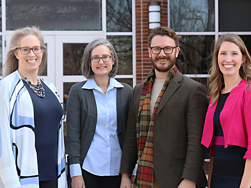 three women and one man standing in a line in front of a building smiling for the camera