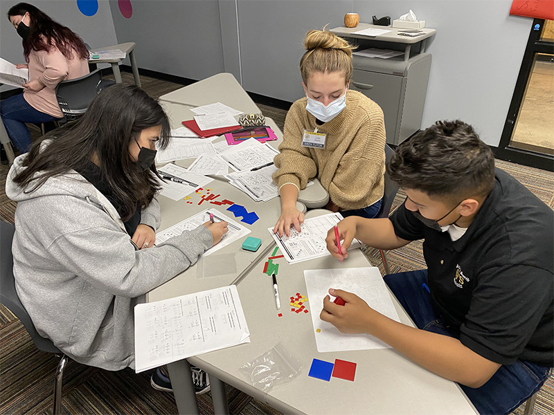 Three students and a tutor in a classroom sitting around a table looking at math worksheets