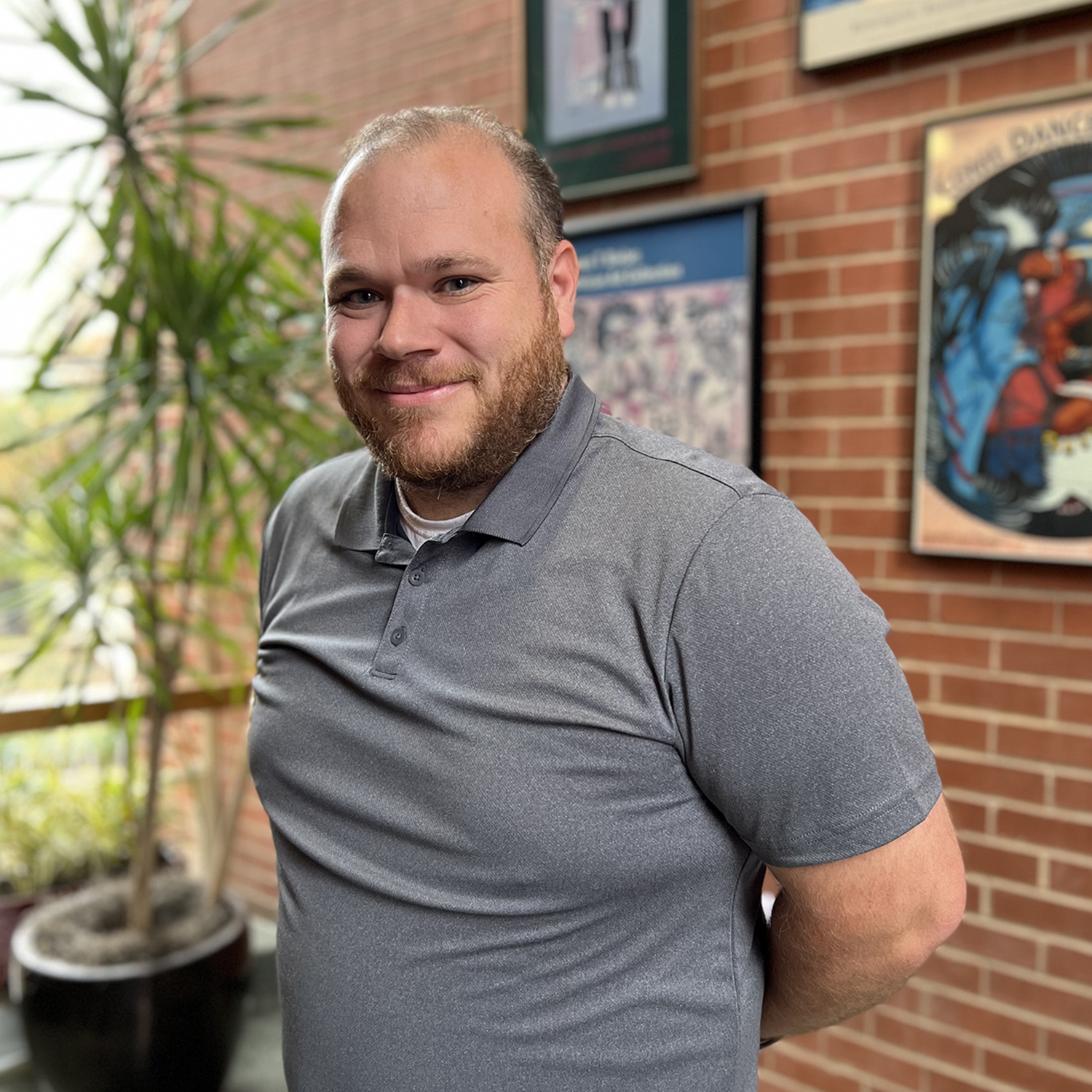 a man in a gray shirt with a beard standing in front of a brick wall