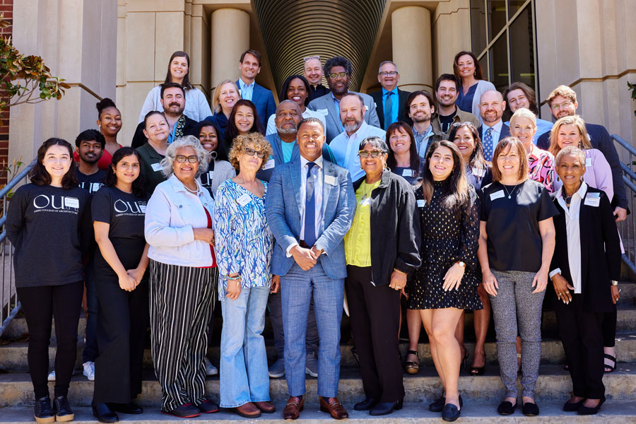 A large group posing for a photo on the steps of the entrance to an academic building.