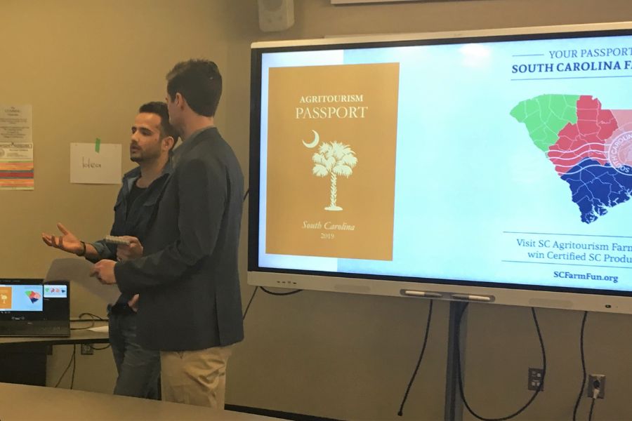 Two students presenting their work in front of a digital board with slide discussing agritourism.