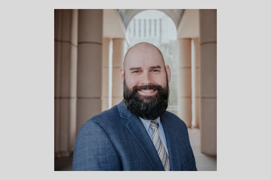 Outdoor headshot between two columns of a bald man with a thick brown bears wearing a tie and a blue sport coat