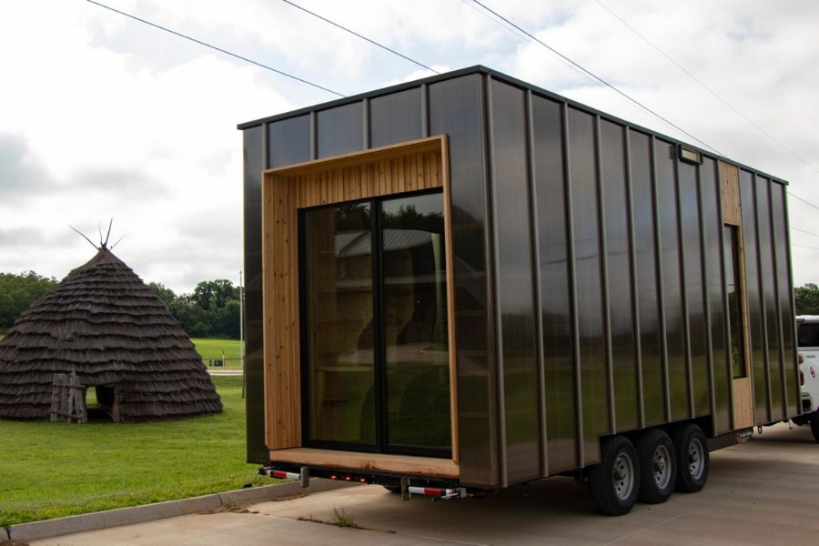 Mobile Med Unit, built on trailer with black metal siding with wood accents, parked near a tribal hut. 