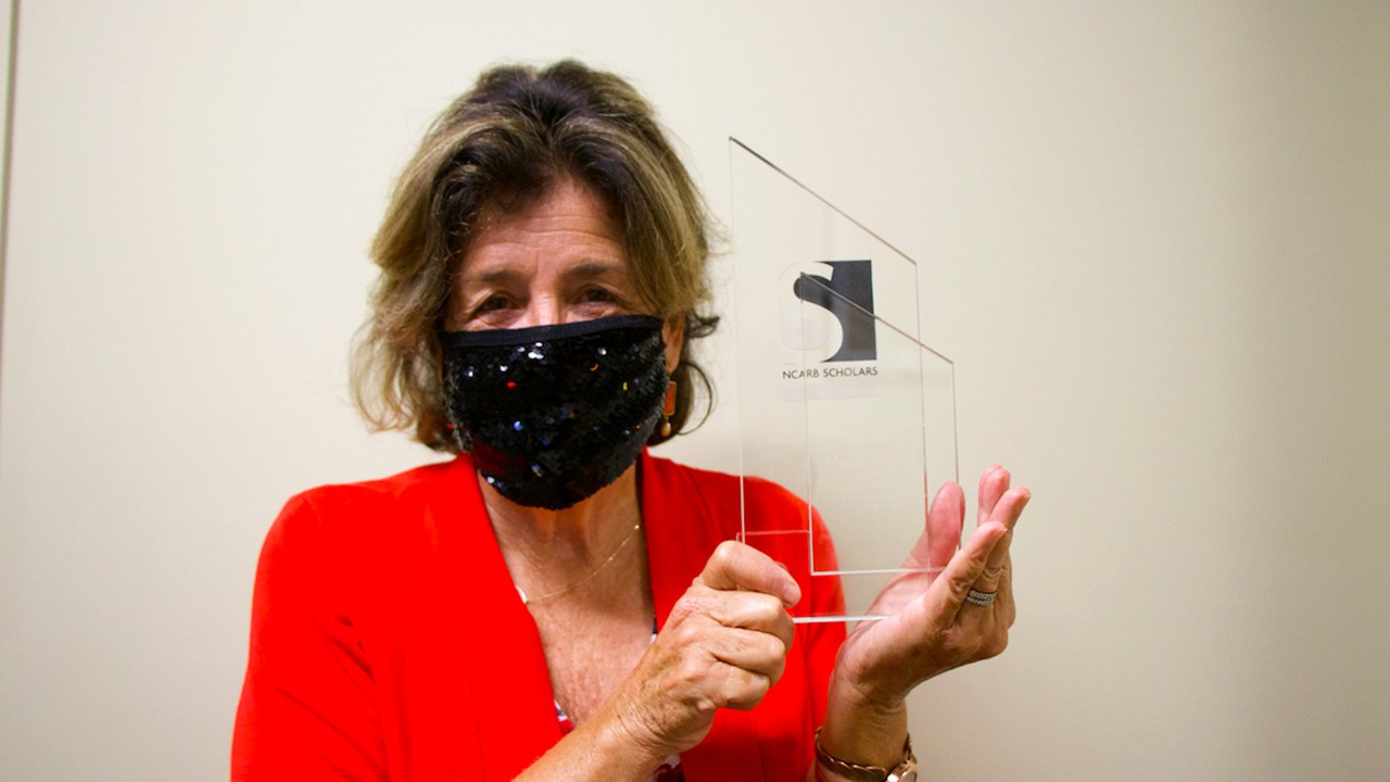 Marjorie Callahan holds her NCARB Scholar award while wearing a face mask and red jacket.