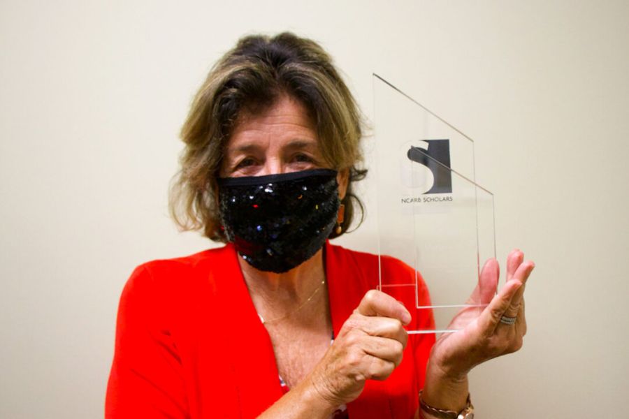 Marjorie Callahan holds her NCARB Scholar award while wearing a face mask and red jacket.