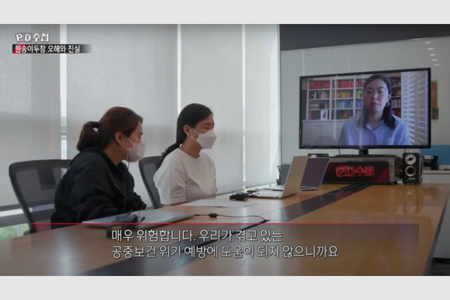 C. Aujean Lee interview with Korean TV outlet over video call, interviewers sitting at conference table and Lee on T V for video call with Korean subtitled text.