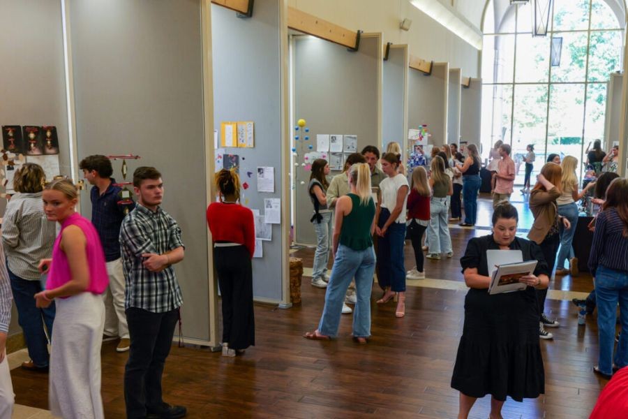 Students mingling and presenting their work, with a professor grading.