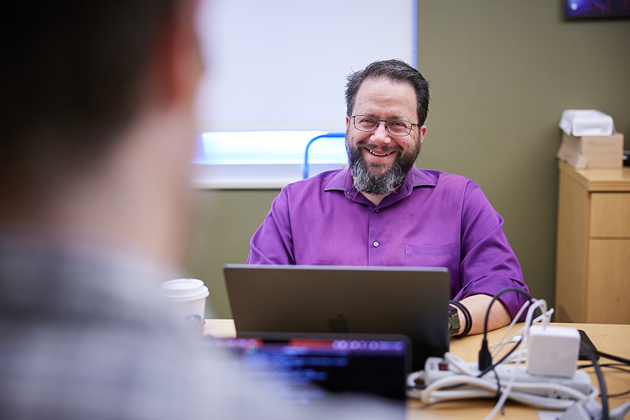 A man in a purple shirt at a computer facing forward and assisting another person.