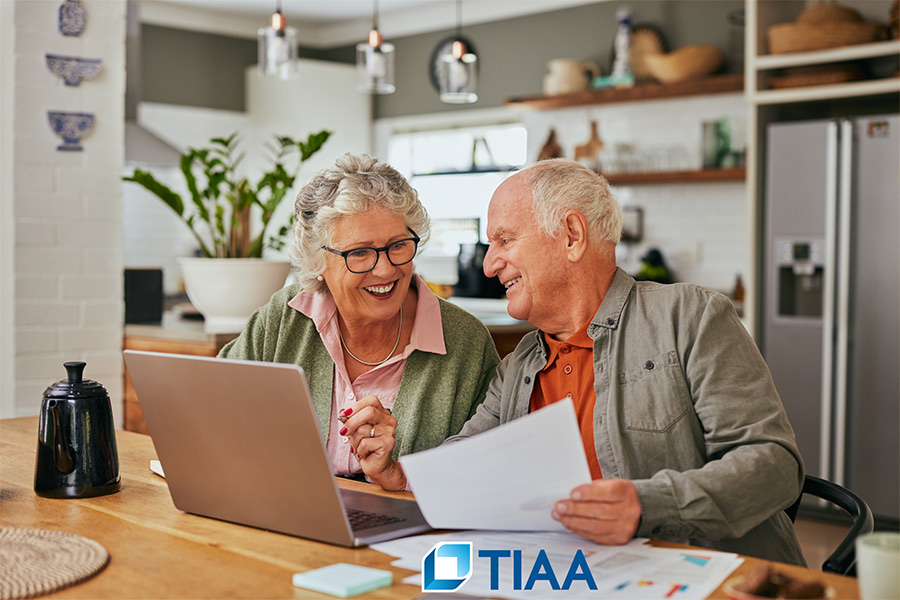 An elderly couple discussing their finances at their dining table with the TIAA logo at the bottom.