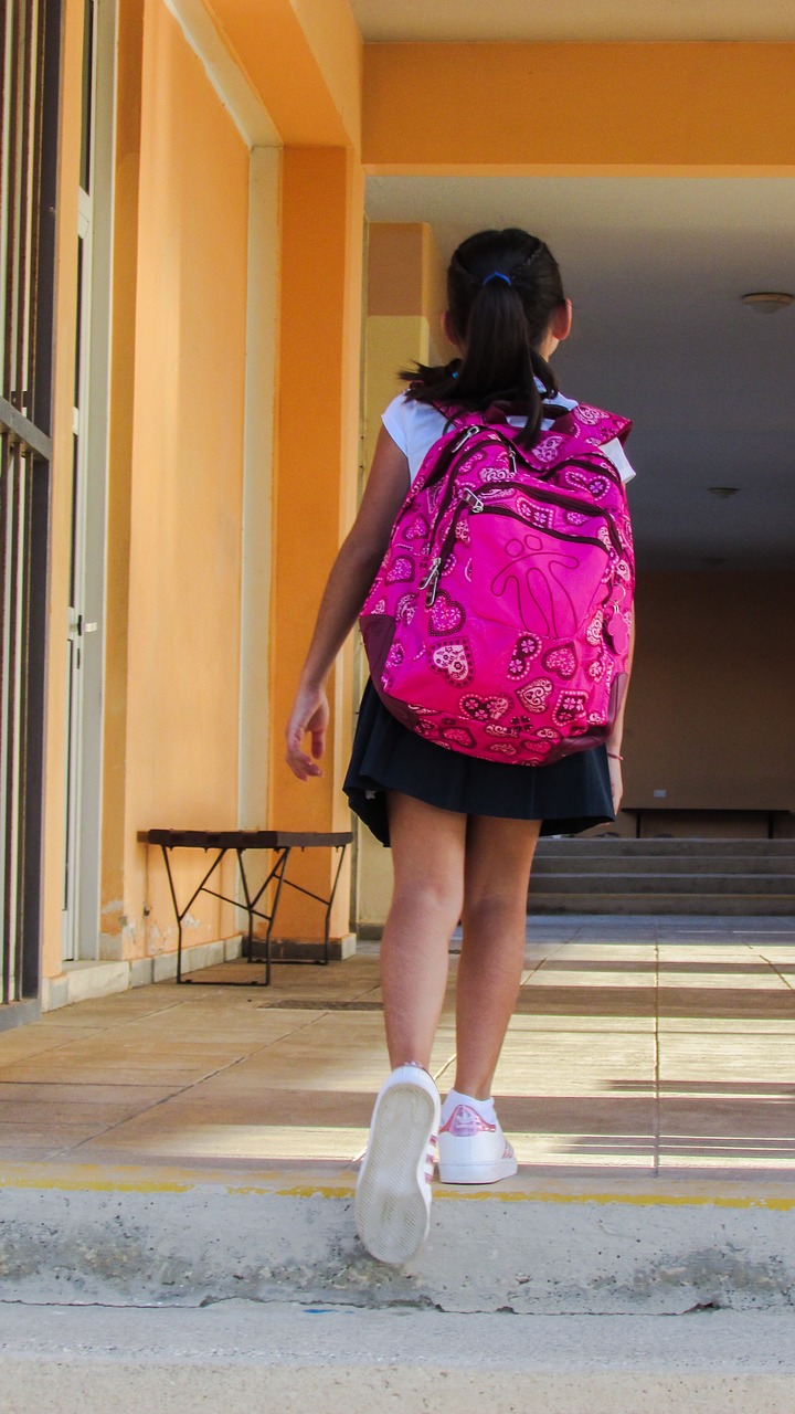 A young girl wearing black skirt and white top is walking away and going up a step with a fuchsia pack on her back. 