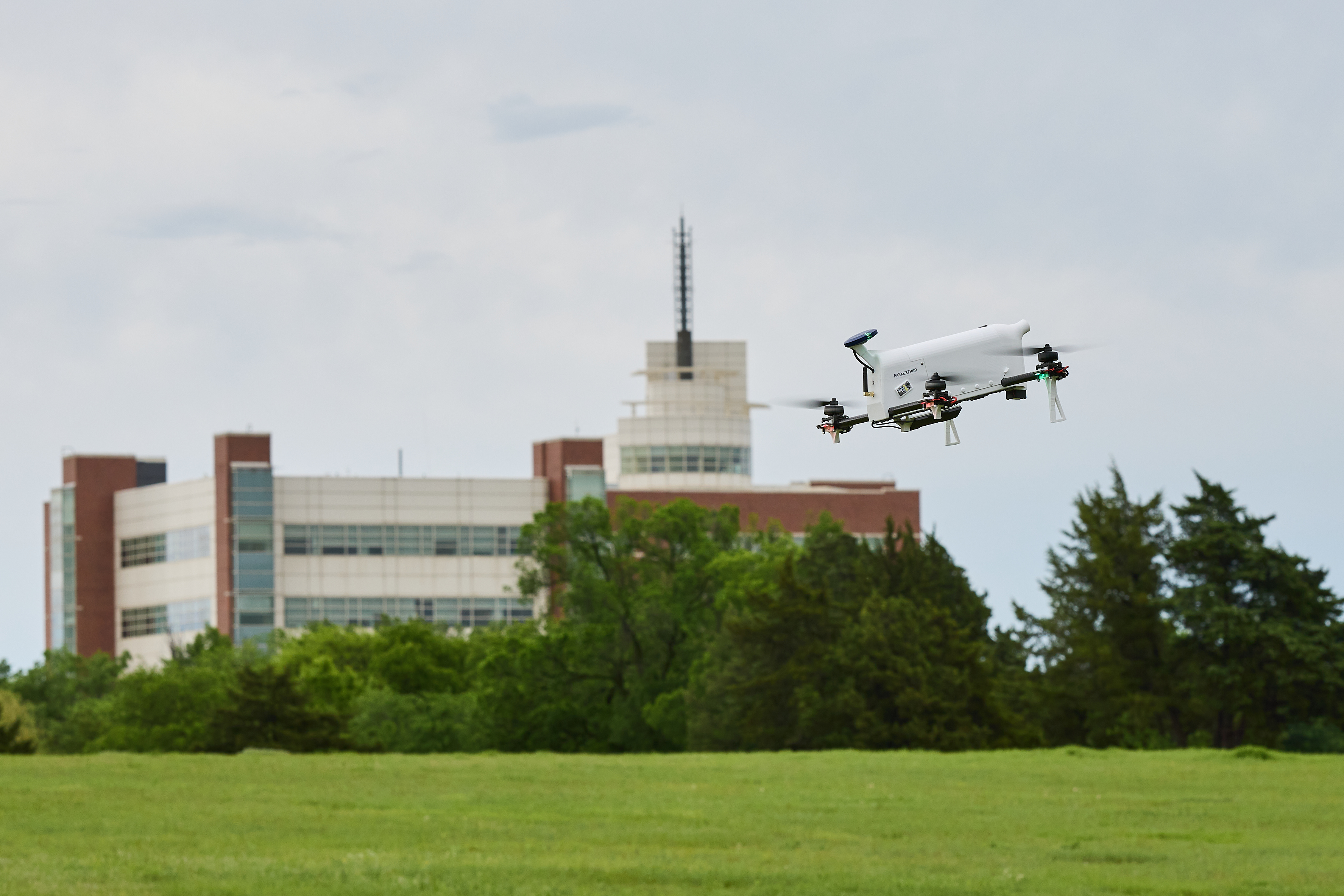 In the foreground, a white copter-style drone ascends. In the background, the National Weather Center sits behind a line of trees.