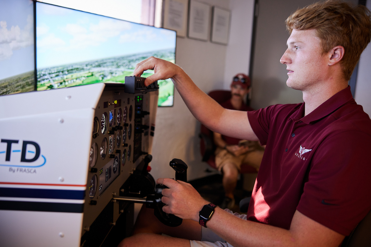 A student in a crimson polo shirt stands in front of an air traffic control simulator.