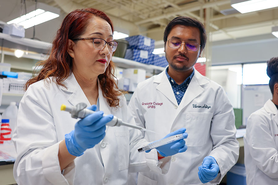 Two researchers in white coats, one holding a pipette, look at a sample. 