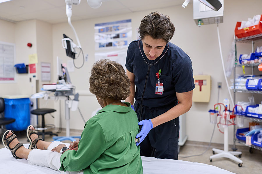 A person in scrubs holds a stethoscope to a woman's back as she sits on a bed in a hospital room.