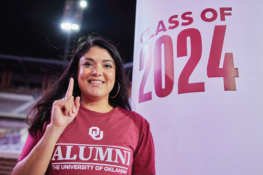 Woman wearing an OU Alumni T-shirt stands in front of a sign that reads "Class of 2024" , She smiles while giving the 'One-OU' hand symbol. 