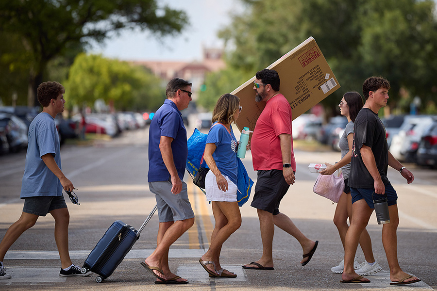 New students and parents on a crosswalk carrying furniture and lugging suitcases on move in day.