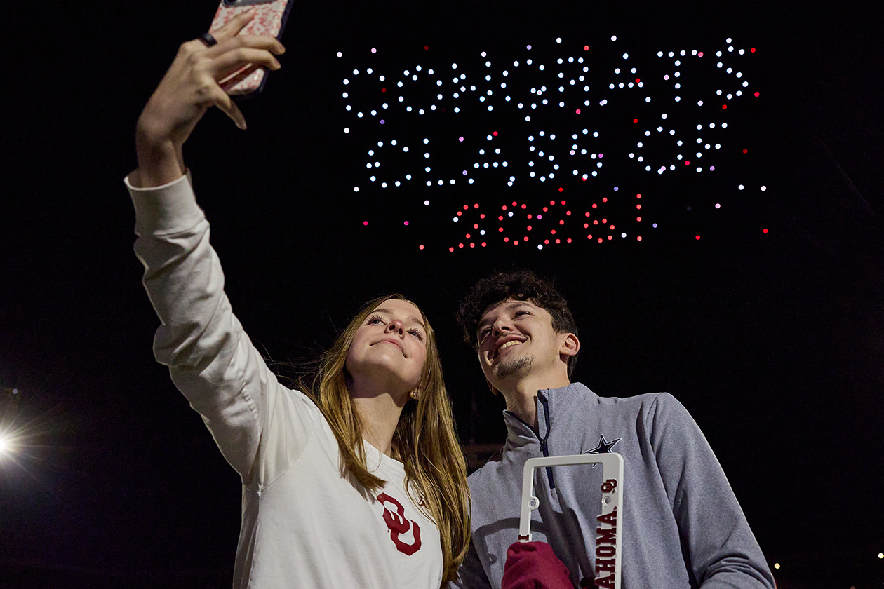 Drones spelling out "Congrats Class of 2026" behind two students taking a selfie.
