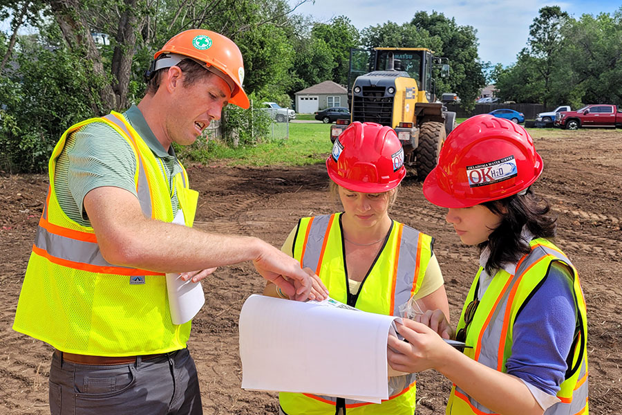 Three people in hard hats look over engineering plans for a site.