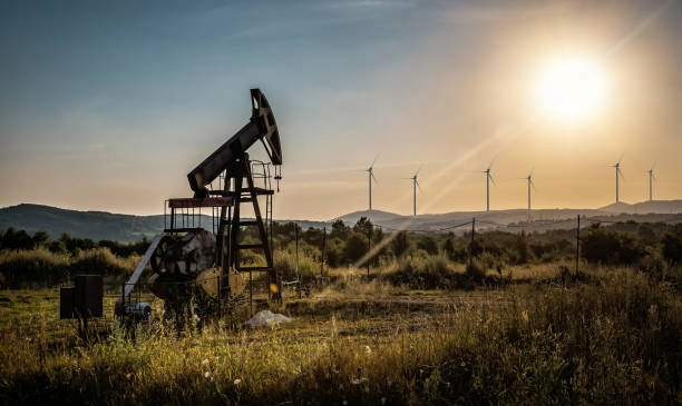 An oil derrick at sunset with wind turbine on a ridgeline in the background