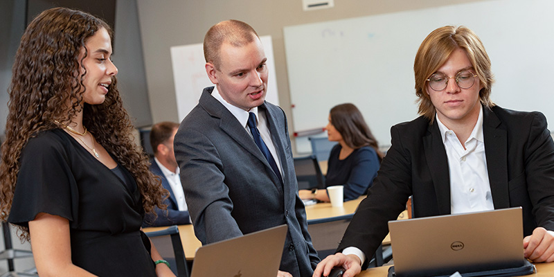 two students work on their laptops as a professor stands nearby to assist