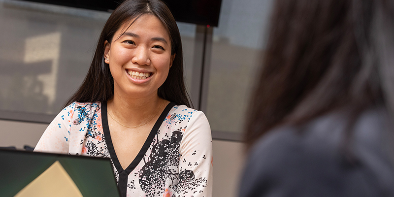 A professionally dressed student smiles at the camera as she looks up from her laptop.