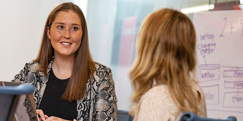 two female students sitting at a table engaged in a one on one conversation.