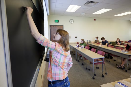 A student writing on a chalkboard in a classroom of students.